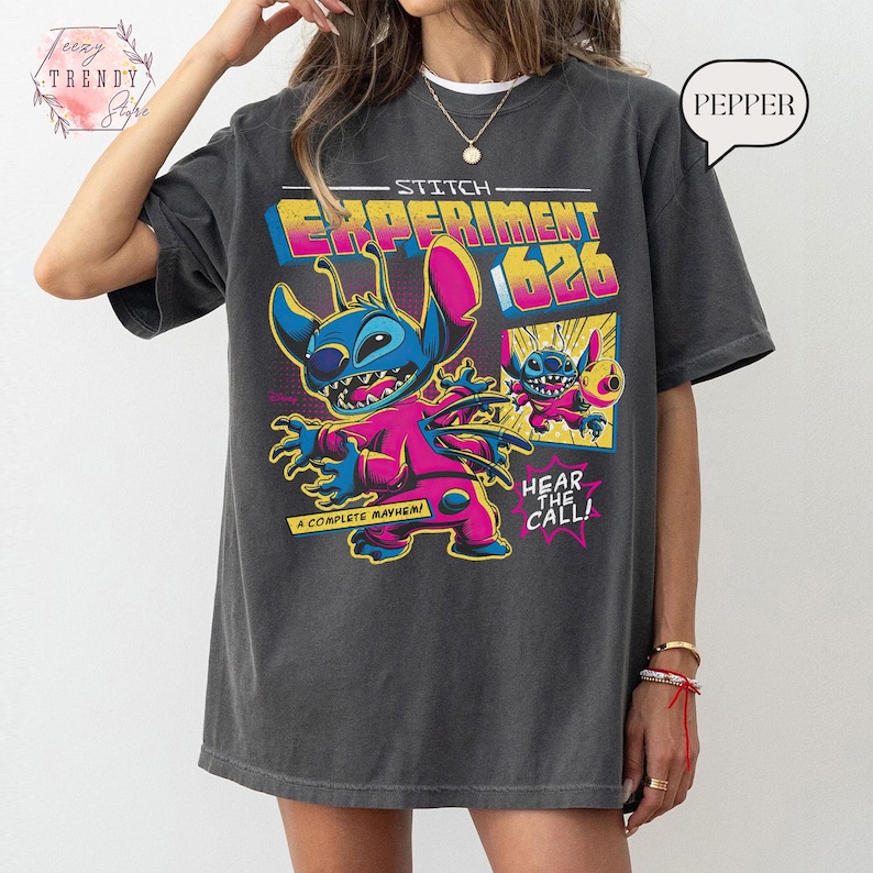 Pode incluir: T-shirt cinza escuro com um gr&aacute;fico colorido de Stitch de Lilo & Stitch. O design inclui o texto "Stitch Experiment 626" e "Hear the Call!" em estilo cartoon. A t-shirt tem um corte folgado.