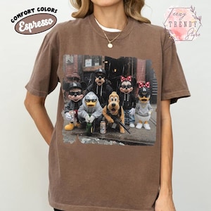 May include: A brown short-sleeve t-shirt featuring a graphic print of five cartoon characters wearing Los Angeles Dodgers baseball caps and casual clothing. The print shows Mickey Mouse, Donald Duck, Goofy, Pluto, and Minnie Mouse in an urban setting.