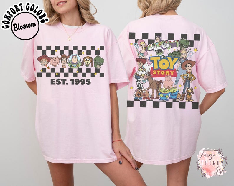 Op de afbeelding: Lichtroze T-shirt met een dambordpatroon en Toy Story-personages, plus de tekst "EST. 1995". De achterkant heeft een grotere Toy Story-afbeelding. Het shirt heeft korte mouwen.