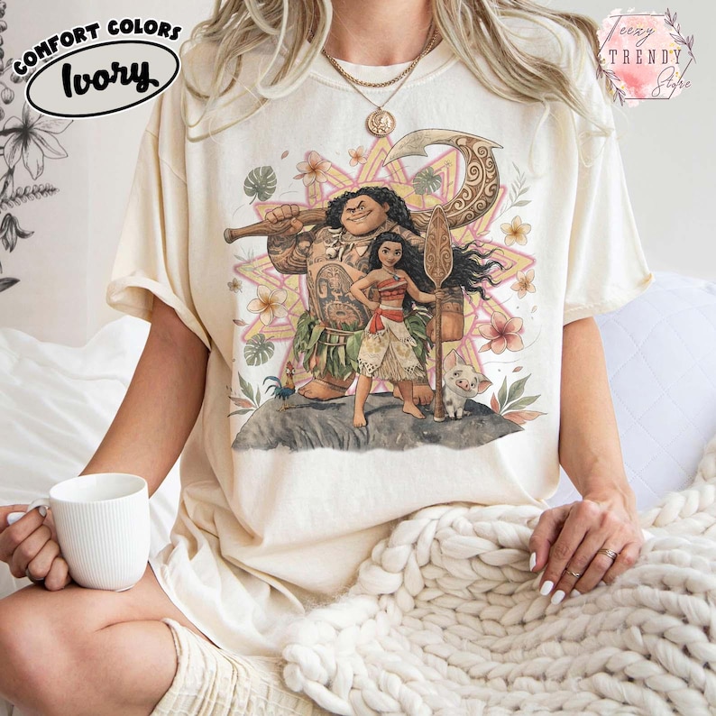 Pu&ograve; includere: T-shirt color avorio con una grafica di personaggi di un film d'animazione. Il design include una grande figura tatuata, una donna con una lancia e un maialino. La maglietta ha le parole "Comfort Colors" e "Ivory".