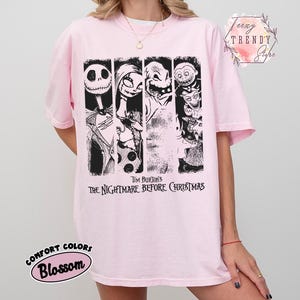 May include: A light pink t-shirt featuring a black and white graphic print of characters from Tim Burton's The Nightmare Before Christmas. The design includes four vertical panels with illustrations of Jack Skellington, Sally, and other figures, with the movie title text printed below.