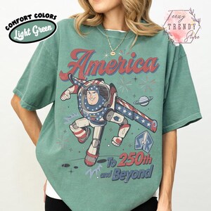 Puede incluir: Una camiseta de cuello redondo verde claro con un gr&aacute;fico de un personaje guardi&aacute;n espacial con un traje patri&oacute;tico. El texto dice America, To 250th and Beyond. El dise&ntilde;o incluye estrellas, rayas, un planeta y fuegos artificiales en rojo, blanco y azul.