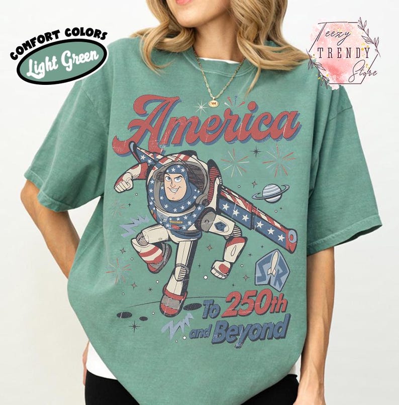 Puede incluir: Una camiseta de cuello redondo verde claro con un gr&aacute;fico de un personaje guardi&aacute;n espacial con un traje patri&oacute;tico. El texto dice America, To 250th and Beyond. El dise&ntilde;o incluye estrellas, rayas, un planeta y fuegos artificiales en rojo, blanco y azul.