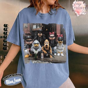 May include: A blue jean colored t-shirt featuring a graphic print of cartoon characters wearing sunglasses, hoodies, and baseball caps. The characters are posed together in an urban setting, with some holding beverage cans and a toy rifle.