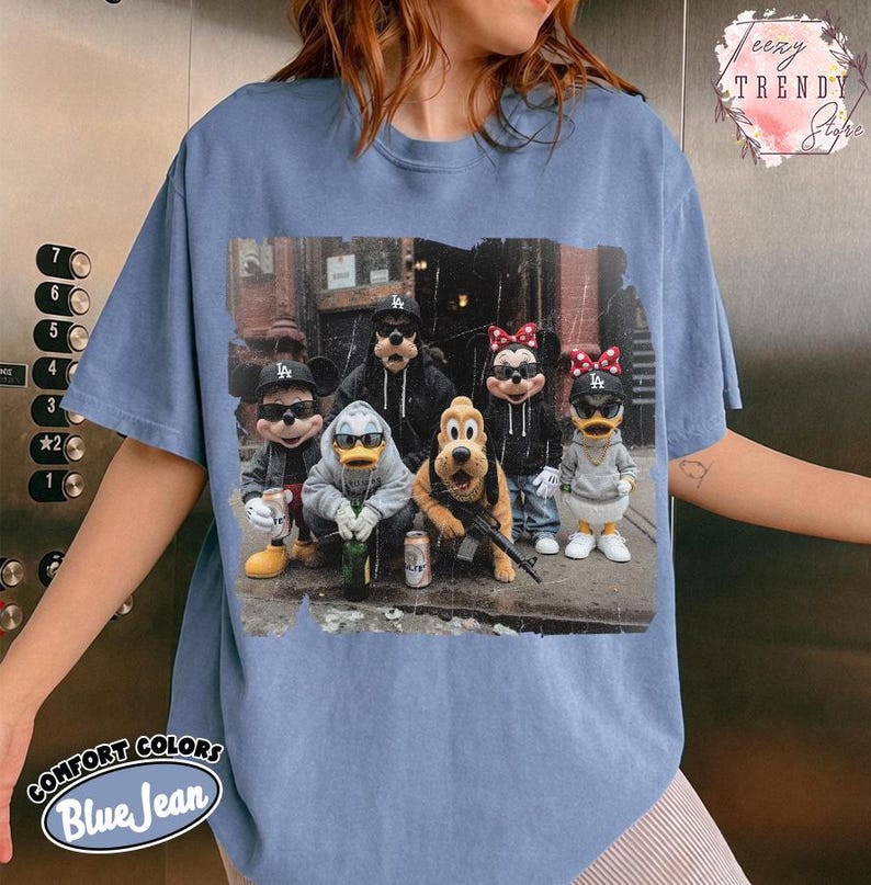 May include: A blue jean colored t-shirt featuring a graphic print of cartoon characters wearing sunglasses, hoodies, and baseball caps. The characters are posed together in an urban setting, with some holding beverage cans and a toy rifle.