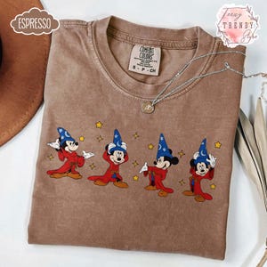 May include: Espresso-colored t-shirt featuring an embroidered design of Mickey Mouse in a wizard costume. The design includes Mickey in a red robe and blue wizard hat with stars. The shirt has a Comfort Colors tag and a silver necklace.