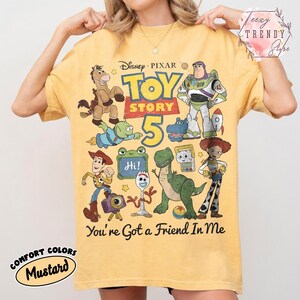 May include: A mustard yellow t-shirt featuring a graphic print of Disney Pixar Toy Story 5 characters including Woody, Buzz Lightyear, Jessie, Rex, and Forky. The design includes the text Toy Story 5 and You've Got a Friend In Me in black.
