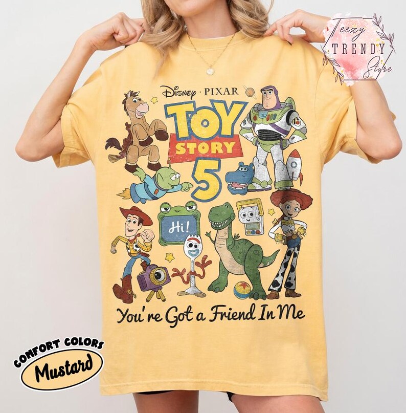 May include: A mustard yellow t-shirt featuring a graphic print of Disney Pixar Toy Story 5 characters including Woody, Buzz Lightyear, Jessie, Rex, and Forky. The design includes the text Toy Story 5 and You've Got a Friend In Me in black.