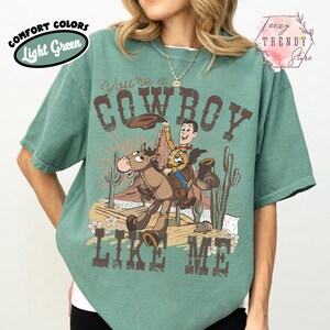 May include: A light green short-sleeve t-shirt featuring a graphic print of a cartoon cowboy riding a horse in a desert landscape. The text reads You're a COWBOY LIKE ME. The design includes cacti, rock formations, and flowers in brown and tan.
