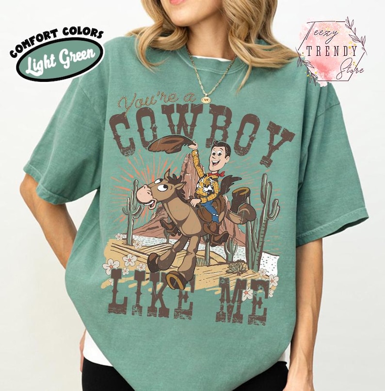 May include: A light green short-sleeve t-shirt featuring a graphic print of a cartoon cowboy riding a horse in a desert landscape. The text reads You're a COWBOY LIKE ME. The design includes cacti, rock formations, and flowers in brown and tan.