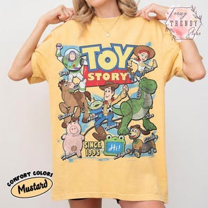 May include: A mustard yellow short-sleeve t-shirt featuring a large graphic print of Toy Story characters including Buzz Lightyear, Woody, Jessie, Bullseye, Rex, Hamm, Slinky, and an Alien. The text reads TOY STORY SINCE 1995 and Hi on a green frog.