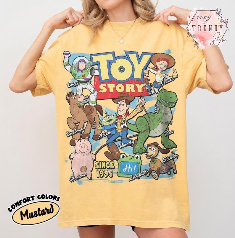 May include: A mustard yellow short-sleeve t-shirt featuring a large graphic print of Toy Story characters including Buzz Lightyear, Woody, Jessie, Bullseye, Rex, Hamm, Slinky, and an Alien. The text reads TOY STORY SINCE 1995 and Hi on a green frog.