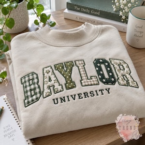 Peut inclure: Un sweat-shirt beige à col rond arborant le mot BAYLOR en grandes lettres appliquées en tissu avec divers motifs verts et beiges, notamment du vichy, des fleurs et des étoiles. Le mot UNIVERSITY est brodé en fil vert foncé.