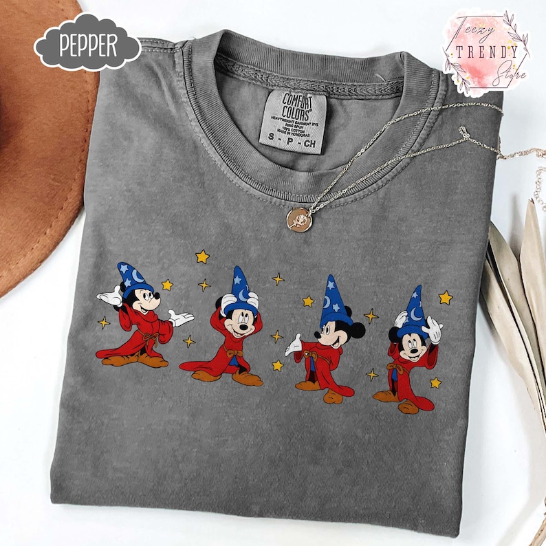May include: A gray Comfort Colors t-shirt featuring a graphic of Mickey Mouse in a sorcerer's outfit. Mickey is wearing a blue hat with stars, a red robe, and is surrounded by yellow stars. A silver necklace with a pendant is draped on the shirt.