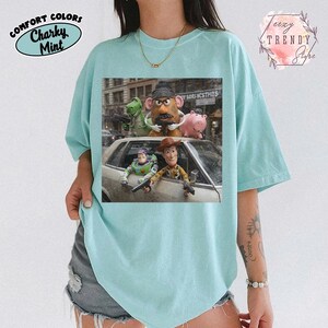 Puede incluir: Una camiseta de color verde menta claro con un estampado gr&aacute;fico de cinco personajes de juguete de una pel&iacute;cula. Los personajes est&aacute;n colocados alrededor de un coche plateado, algunos sosteniendo peque&ntilde;as pistolas de juguete negras. El estampado incluye un dinosaurio verde, una figura con forma de patata, un cerdo rosa, un guardi&aacute;n espacial y un vaquero.