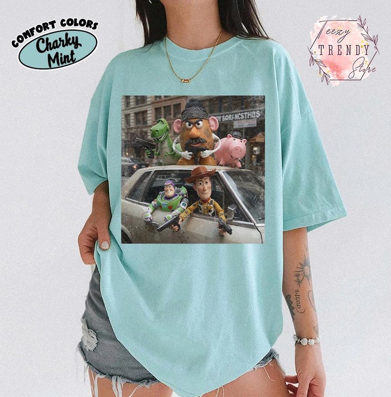Puede incluir: Una camiseta de color verde menta claro con un estampado gr&aacute;fico de cinco personajes de juguete de una pel&iacute;cula. Los personajes est&aacute;n colocados alrededor de un coche plateado, algunos sosteniendo peque&ntilde;as pistolas de juguete negras. El estampado incluye un dinosaurio verde, una figura con forma de patata, un cerdo rosa, un guardi&aacute;n espacial y un vaquero.