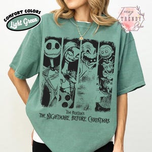 May include: A light green short-sleeve t-shirt featuring a black graphic print of characters from Tim Burton's The Nightmare Before Christmas. The design includes Jack Skellington, Sally, Oogie Boogie, and other figures arranged in vertical panels.