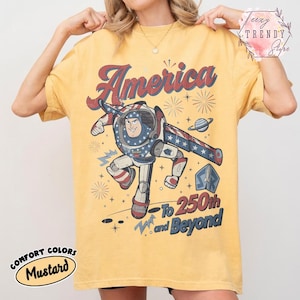 Puede incluir: Una camiseta de color amarillo mostaza con un gr&aacute;fico de un personaje de guardi&aacute;n espacial con traje rojo, blanco y azul. El texto dice America, To 250th and Beyond, con ilustraciones de fuegos artificiales, estrellas, un planeta y un cohete.