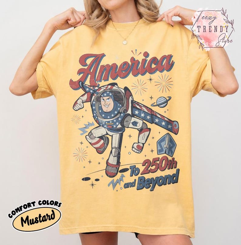 Puede incluir: Una camiseta de color amarillo mostaza con un gr&aacute;fico de un personaje de guardi&aacute;n espacial con traje rojo, blanco y azul. El texto dice America, To 250th and Beyond, con ilustraciones de fuegos artificiales, estrellas, un planeta y un cohete.