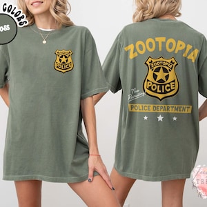 May include: Moss green t-shirt featuring a gold and black police badge graphic on the front and back. The back of the shirt displays the text "ZOOTOPIA POLICE DEPARTMENT" in yellow. The shirt is a short-sleeved, unisex style.