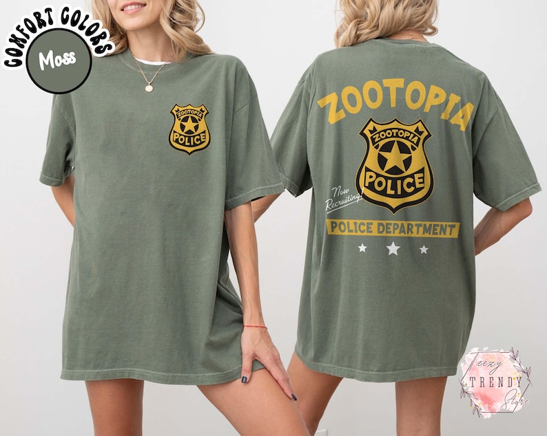May include: Moss green t-shirt featuring a gold and black police badge graphic on the front and back. The back of the shirt displays the text "ZOOTOPIA POLICE DEPARTMENT" in yellow. The shirt is a short-sleeved, unisex style.