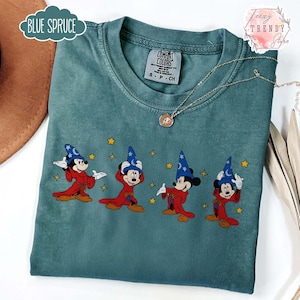 May include: Blue Spruce Comfort Colors t-shirt featuring a graphic of Mickey Mouse in a wizard costume. The design includes Mickey in a red robe and blue wizard hat with stars. The shirt has a crew neck and short sleeves.