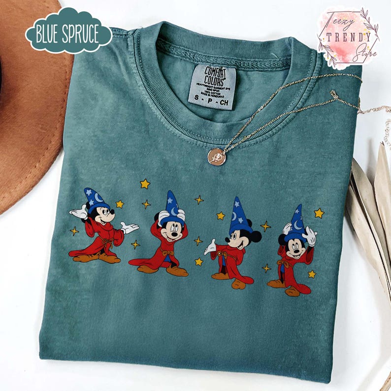 May include: Blue Spruce Comfort Colors t-shirt featuring a graphic of Mickey Mouse in a wizard costume. The design includes Mickey in a red robe and blue wizard hat with stars. The shirt has a crew neck and short sleeves.
