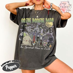 May include: A charcoal gray short-sleeve t-shirt featuring a graphic print with the text Oogie Boogie Bash and The Spookiest Place on Earth. The design includes illustrations of characters, a haunted mansion, a moon, and fireworks.
