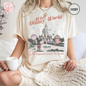 May include: Ivory-colored t-shirt featuring a vintage-style graphic of Walt Disney World. The design includes Cinderella's castle, Mickey Mouse, and the text "The happiest place on earth."