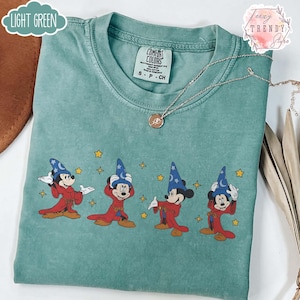 May include: Light green Comfort Colors t-shirt featuring a graphic of Mickey Mouse in a sorcerer's outfit. The design shows Mickey in various poses with a blue wizard hat and red robe. A silver necklace with a pendant is also visible.