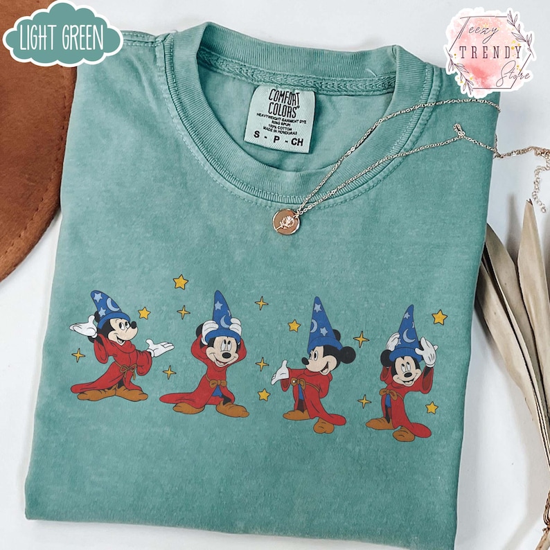 May include: Light green Comfort Colors t-shirt featuring a graphic of Mickey Mouse in a sorcerer's outfit. The design shows Mickey in various poses with a blue wizard hat and red robe. A silver necklace with a pendant is also visible.