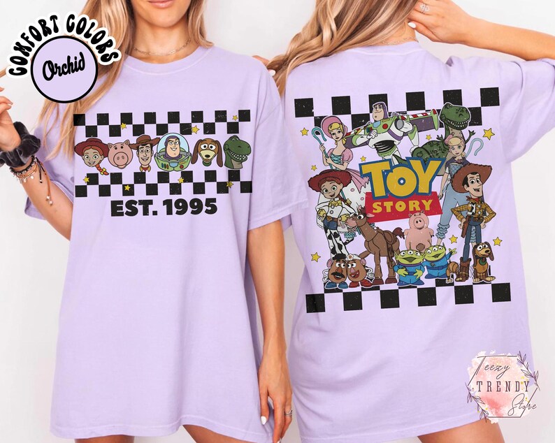 Op de afbeelding: Een lichtpaars T-shirt met een dambordpatroon en stripfiguren, met de tekst "EST. 1995". De achterkant van het shirt toont een "Toy Story"-afbeelding. Het T-shirt heeft een ronde hals en korte mouwen.
