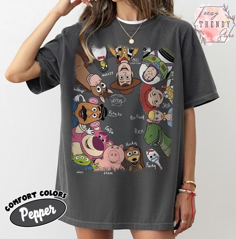 May include: Dark gray t-shirt featuring a colorful graphic of Toy Story characters, including Woody, Buzz Lightyear, and Rex. The shirt has the words "COMFORT COLORS Pepper" printed on the bottom. The shirt is a crew neck.