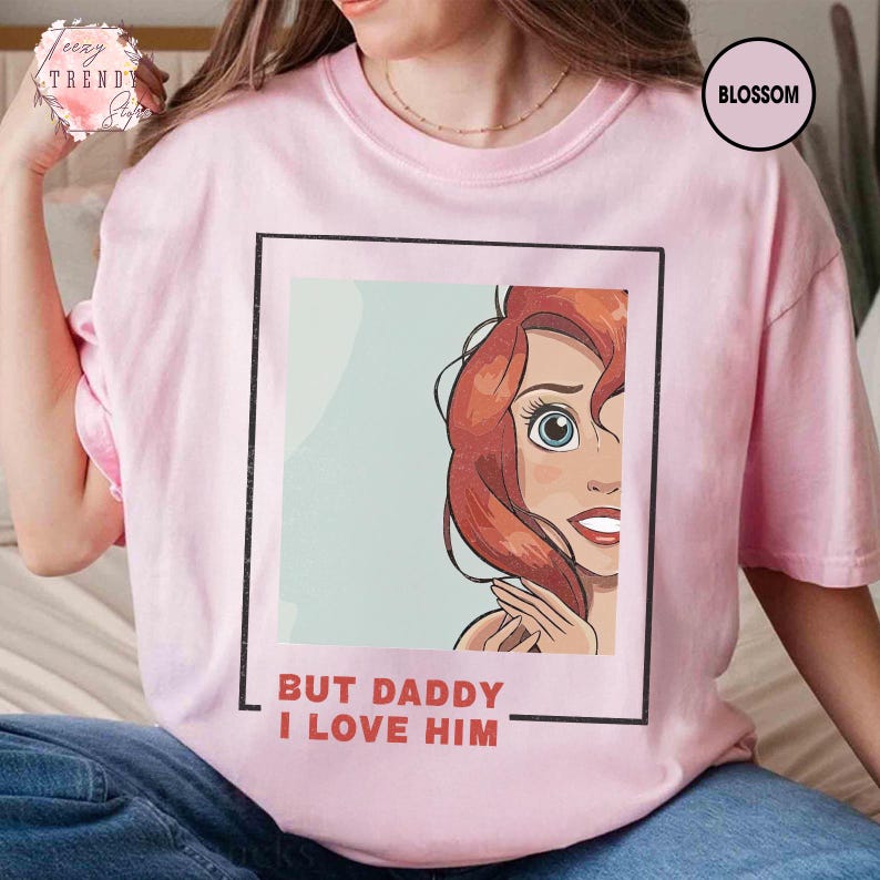 Puede incluir: Camiseta rosa claro con una ilustraci&oacute;n de dibujos animados de un personaje con cabello rojo y ojos azules. El texto "BUT DADDY I LOVE HIM" est&aacute; impreso en rojo. La camiseta tiene cuello redondo y mangas cortas.