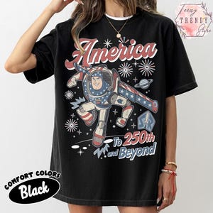 Puede incluir: Una camiseta negra de manga corta con un gr&aacute;fico de un personaje de guardi&aacute;n espacial con un traje rojo, blanco y azul. El dise&ntilde;o incluye el texto America, 250th y Beyond, junto con ilustraciones de estrellas y fuegos artificiales al frente.