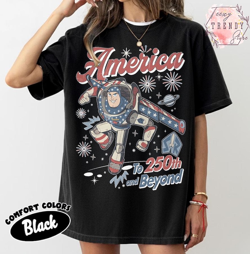 Puede incluir: Una camiseta negra de manga corta con un gr&aacute;fico de un personaje de guardi&aacute;n espacial con un traje rojo, blanco y azul. El dise&ntilde;o incluye el texto America, 250th y Beyond, junto con ilustraciones de estrellas y fuegos artificiales al frente.