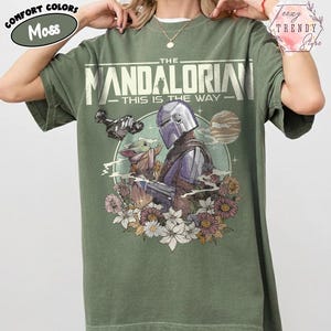 May include: A moss green t-shirt with a graphic of The Mandalorian and Grogu, encircled by flowers. The text reads "THE MANDALORIAN - THIS IS THE WAY". The shirt has a loose fit and is made of a soft material.