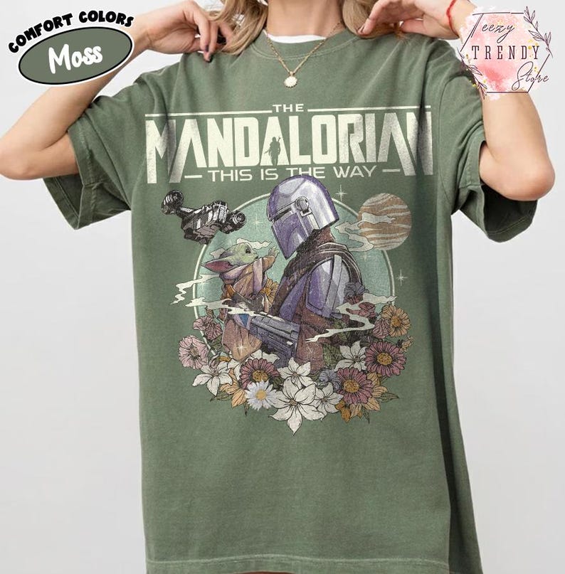 May include: A moss green t-shirt with a graphic of The Mandalorian and Grogu, encircled by flowers. The text reads "THE MANDALORIAN - THIS IS THE WAY". The shirt has a loose fit and is made of a soft material.