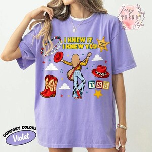 May include: A violet t-shirt featuring a graphic print with the text I KNEW IT, I KNEW YOU, a cartoon character playing a guitar, red cowboy boots, a red hat, a sheriff star badge, and colourful blocks with the letters TS5 on a light background.