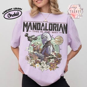 May include: A lilac t-shirt featuring a graphic of The Mandalorian and Grogu, with floral elements. The text "THE MANDALORIAN" and "THIS IS THE WAY" are printed on the front. The shirt is labelled "Comfort Colors" and "Orchid".
