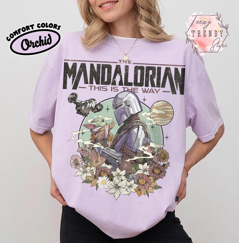 May include: A lilac t-shirt featuring a graphic of The Mandalorian and Grogu, with floral elements. The text "THE MANDALORIAN" and "THIS IS THE WAY" are printed on the front. The shirt is labelled "Comfort Colors" and "Orchid".