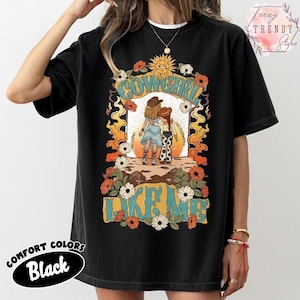 May include: A black t-shirt featuring a graphic print of two figures in cowboy hats and western attire standing together. The design includes a sun, flowers, flames, and the text COWGIRL LIKE ME in a retro font style. The shirt has a crew neckline.