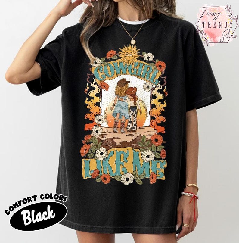 May include: A black t-shirt featuring a graphic print of two figures in cowboy hats and western attire standing together. The design includes a sun, flowers, flames, and the text COWGIRL LIKE ME in a retro font style. The shirt has a crew neckline.