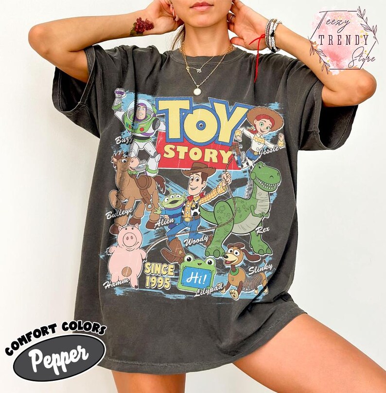May include: A charcoal gray short-sleeve t-shirt featuring a graphic print of Toy Story characters including Woody, Buzz Lightyear, Jessie, Rex, Bullseye, Hamm, Slinky, and an Alien. The text reads Toy Story, Since 1995, and Hi! Lilypad.