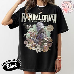 May include: Black t-shirt with a graphic of The Mandalorian and Grogu, floral elements, and the text "THE MANDALORIAN - THIS IS THE WAY." Made from Comfort Colors material.