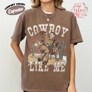 May include: An espresso brown short-sleeve t-shirt featuring a graphic print of a cartoon cowboy riding a horse in a desert landscape with cacti. The text "COWBOY LIKE ME" is printed in a distressed white font above and below the illustration.