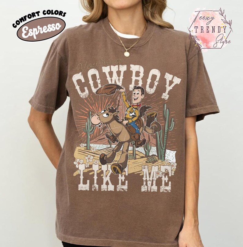 May include: An espresso brown short-sleeve t-shirt featuring a graphic print of a cartoon cowboy riding a horse in a desert landscape with cacti. The text "COWBOY LIKE ME" is printed in a distressed white font above and below the illustration.