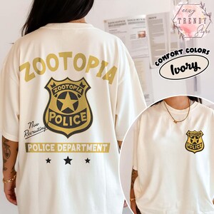 May include: Ivory-colored t-shirt featuring the word "ZOOTOPIA" above a police badge graphic. The badge includes the text "ZOOTOPIA POLICE" and a star. Below the badge is "POLICE DEPARTMENT" and three stars. The shirt also has the text "Now Recruiting".
