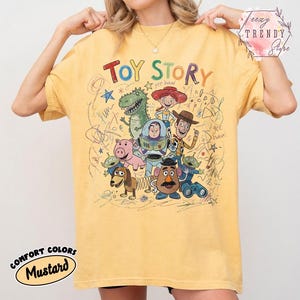 May include: Mustard yellow t-shirt featuring a colorful graphic of Toy Story characters. The text "Toy Story" is above the characters. The shirt has a crew neck and short sleeves. The words "Comfort Colors" and "Mustard" are printed on the bottom left.