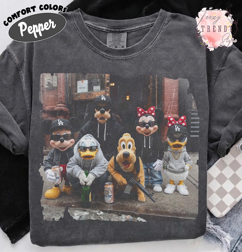 May include: A charcoal gray crewneck t-shirt featuring a graphic print of cartoon characters wearing black baseball caps, sunglasses, and hoodies. The characters are posed in an urban setting, with some holding bottles and one holding a rifle.